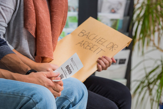 Bachelorarbeit Zwei Personen sitzen nebeneinander. Man sieht einen Ausschnitt von Knie bis Brust. Die hintere Person hat einen großen Umschlag mit der Aufschrift „Bachelor-Arbeit“ in der Hand, die vordere Person hat einen kleinen weißen Zettel mit einer Wartenummer, einem Code und dem TU-Logo in der Hand.